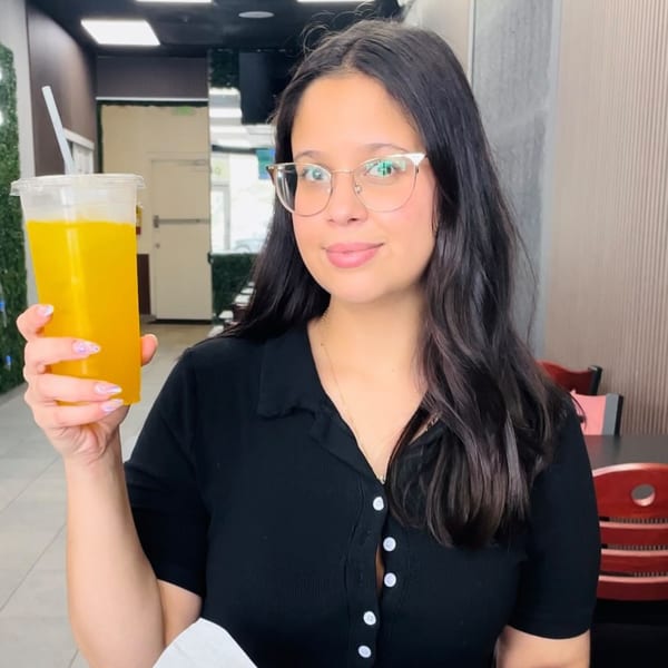 A woman in glasses smiles, holding up a large clear cup of yellow juice with a straw.