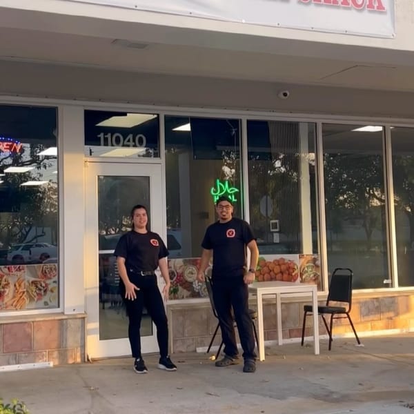 Two Shawarma Shack employees in black shirts stand outside the restaurant with a green Halal sign.