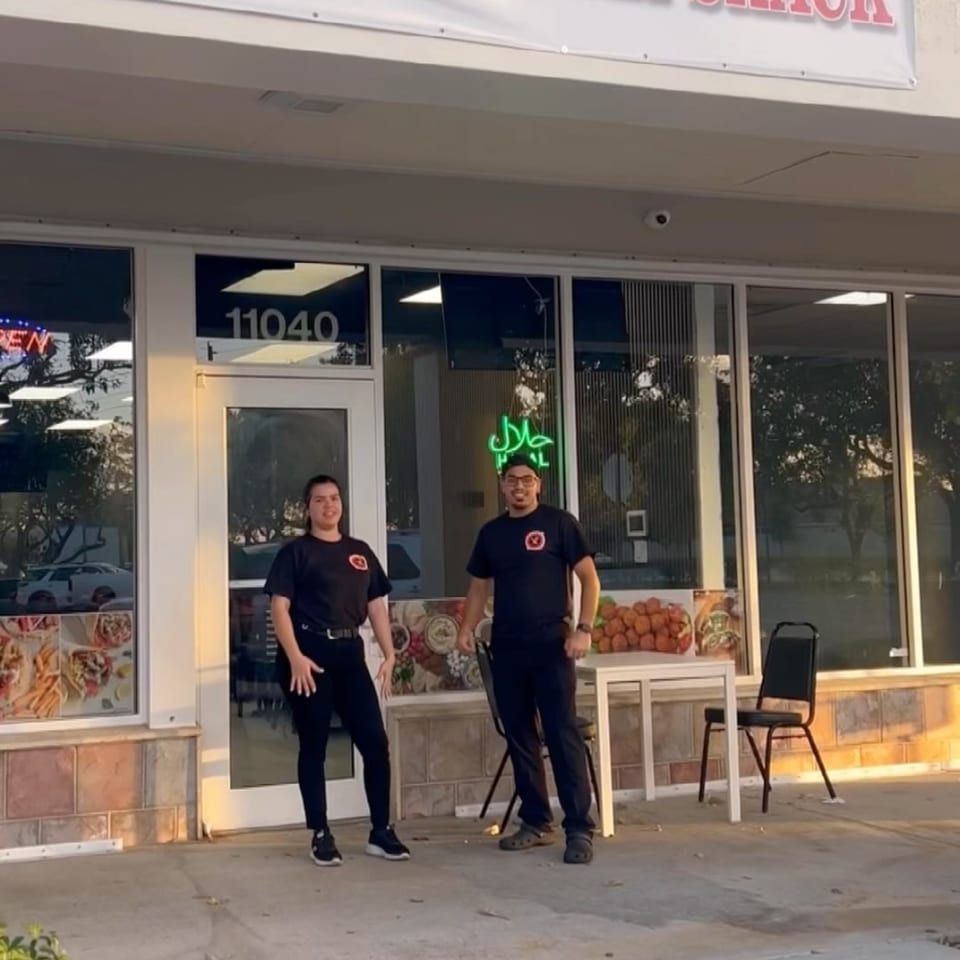 Two Shawarma Shack employees in black shirts stand outside the restaurant with a green Halal sign.