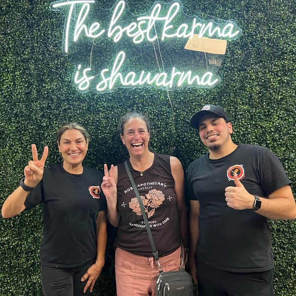 Three smiling people pose in front of a green wall with a neon sign reading "The best karma is shawarma".