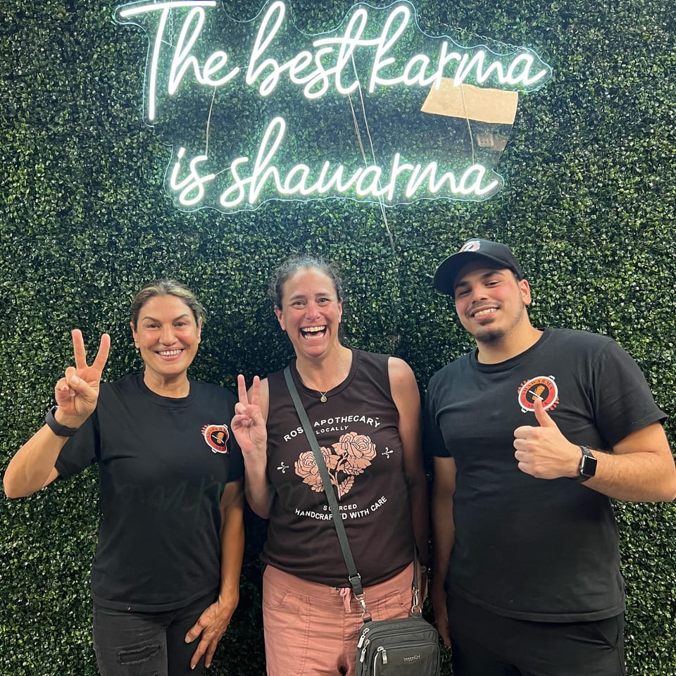 Three smiling people pose in front of a green wall with a neon sign reading "The best karma is shawarma".
