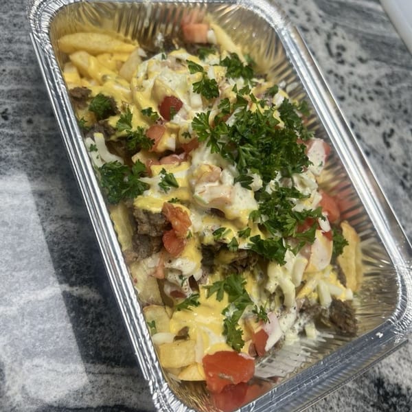 Loaded fries with meat, cheese sauce, white sauce, diced tomatoes, onions, and parsley in a foil container.