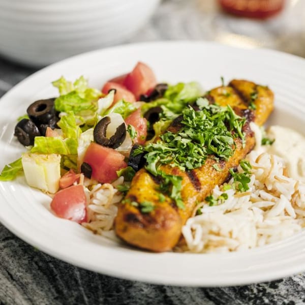 Grilled chicken skewer with rice, a side salad of lettuce, tomatoes, and olives, and a creamy sauce.